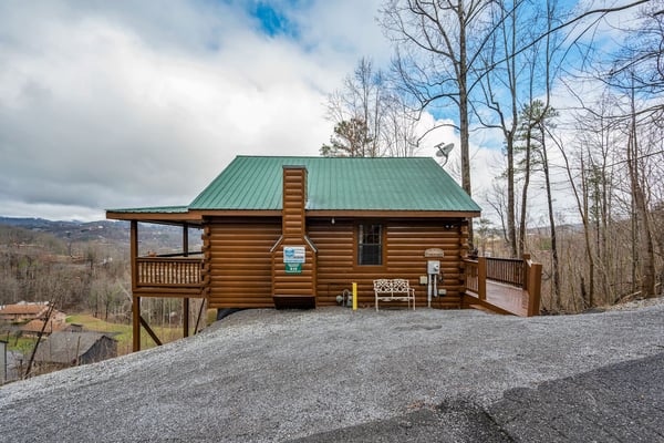 Small wooden cabin with metal roof and front porch, surrounded by green hills and larger cabin in background